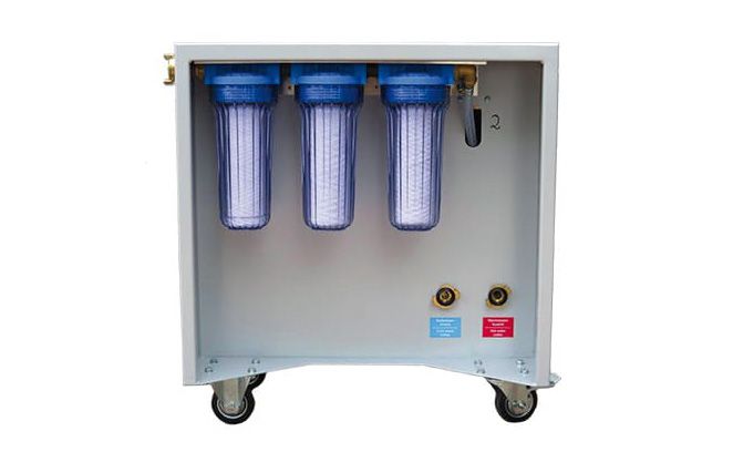 Eine Wasserfiltereinheit mit drei transparenten Filterbehältern, die blau gefärbt sind. Die Einheit hat Räder für die Mobilität und ist mit zwei Anschlüssen in rot und blau ausgestattet, die möglicherweise für Ein- und Auslauf dienen.