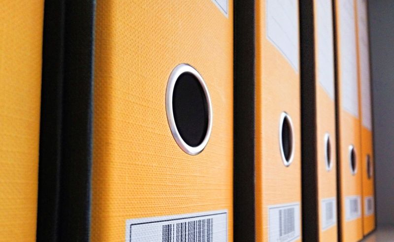 Eine Reihe von orangefarbenen Ordnern steht nebeneinander, wobei jeder Ordner ein rundes Loch in der Mitte hat. Die Ordner sind leicht schräg angeordnet, wodurch die Struktur und Textur der Oberfläche sichtbar wird.