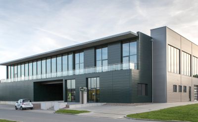 Ein modernes, mehrstöckiges Gebäude mit großen Fenstern und einem flachen Dach. Die Fassade ist in dunklem Grün gehalten. Vor dem Gebäude steht ein Parkplatz. Es scheint in einem Gewerbegebiet zu liegen, umgeben von Rasenflächen.