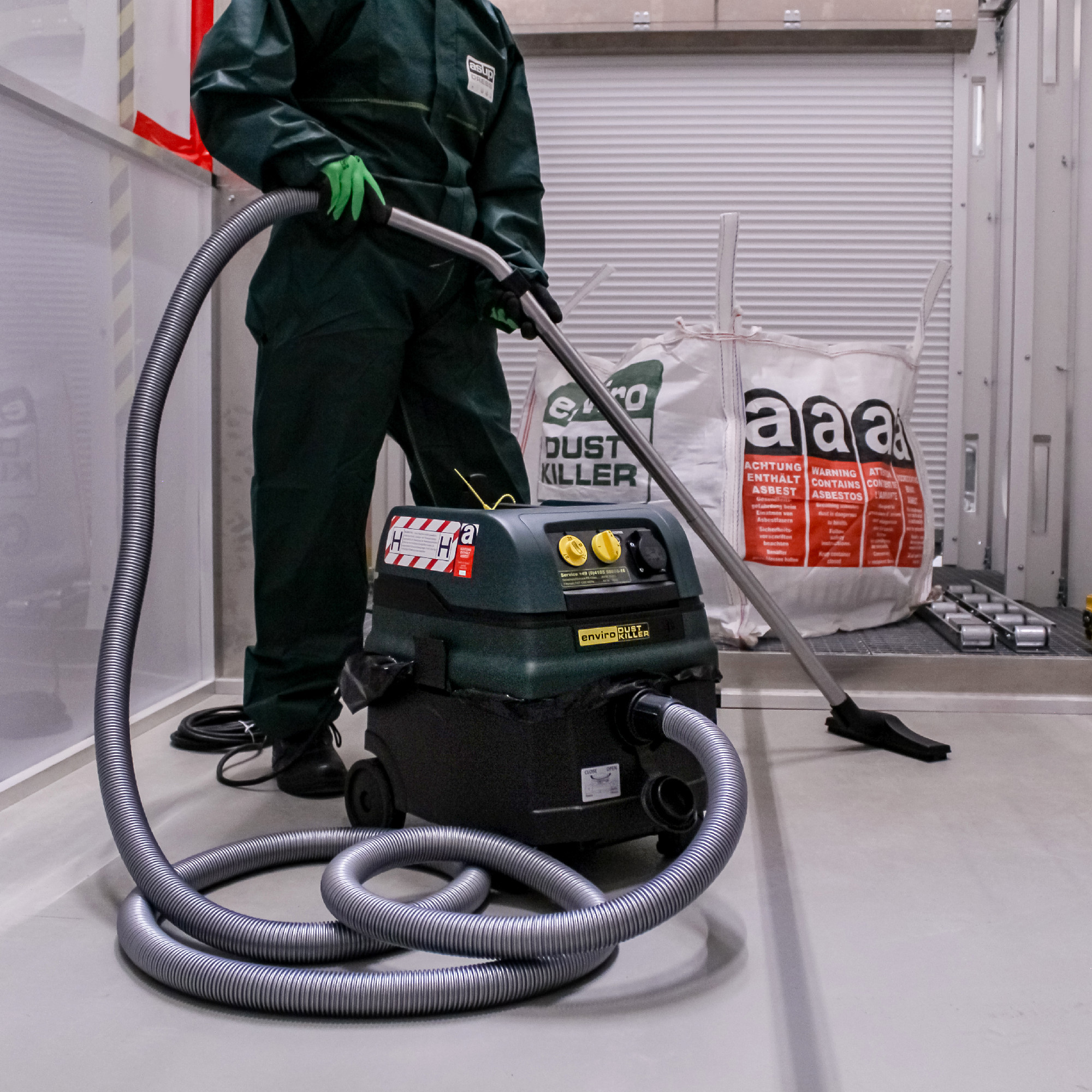 Eine Person in einem grünen Overall steht neben einem Staubsauger mit Schlauch. Im Hintergrund sind Säcke mit der Aufschrift „DUST KILLER“ und Warnhinweise zu Asbest sichtbar. Das Bild vermittelt den Eindruck einer Reinigungsumgebung, die möglicherweise besondere Vorsichtsmaßnahmen erfordert.